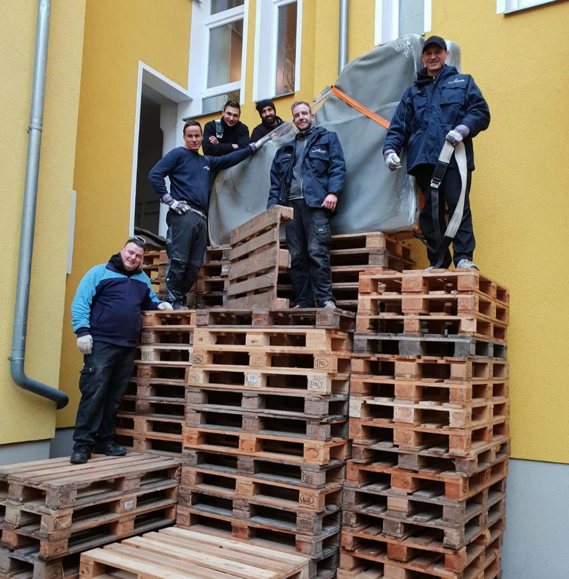 Piano-Express team carrying a packed grand piano over pallet steps in a courtyard